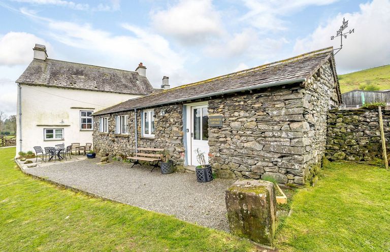 An exterior view of a stone and white painted cottage with outdoor seating on a gravel patio at Shepherds Cottage in Hawkshead