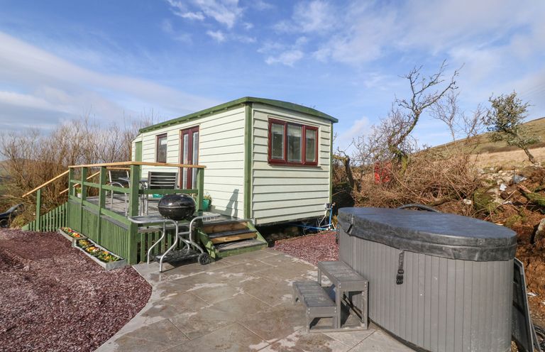 An outdoor area with a small green cabin with a deck and outdoor furniture a charcoal grill and a covered hot tub at Nyth Y Bioden Magpies Nest Llanaelhaearn near Trefor