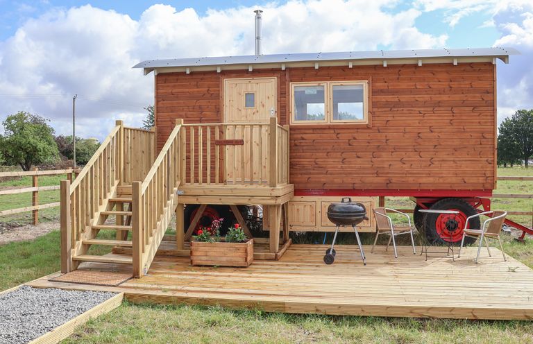 A wooden shepherds hut on wheels with wooden stairs and a small deck with a barbecue grill two chairs and a table at Shepherds Hut in Garthorpe near Scunthorpe