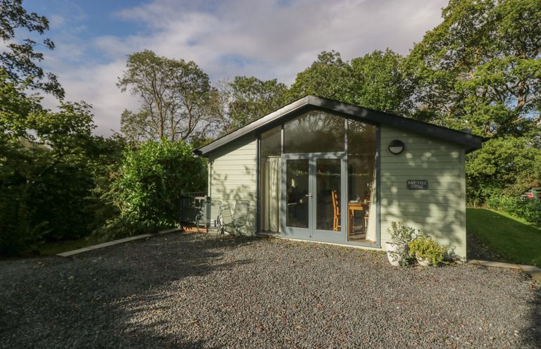 A small lodge with glass doors and chairs outside surrounded by trees at Bay Tree Lodge in Cartmel