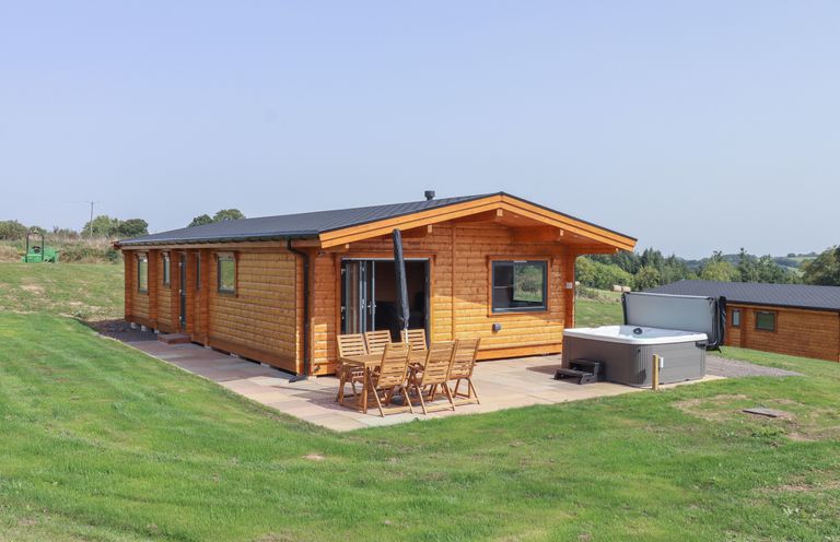 A wooden lodge with patio furniture and a hot tub on a grassy lawn at Foxglove Lodge in Hittisleigh