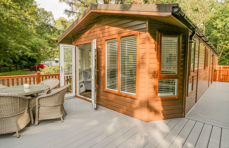 A wooden cabin with glass doors open to a deck with wicker chairs and a glass table surrounded by trees at Willows Lodge White Cross Bay near Windermere