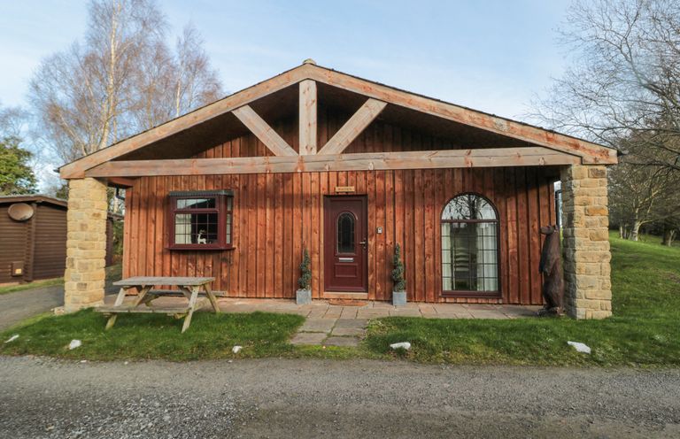 A wooden cabin with a picnic table outside and trees in the background at Matfen in Longframlington