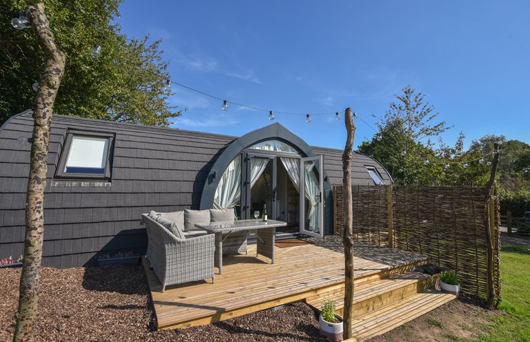 An outdoor wooden deck with seating and table in front of a pod-style building with string lights and trees at Maple in Wootton Fitzpaine near Charmouth