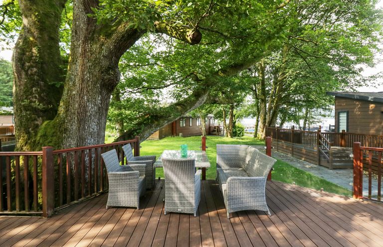 An outdoor wooden deck with wicker chairs and a table surrounded by trees at Tranquil Waters in Lakeside 16 White Cross Bay near Windermere