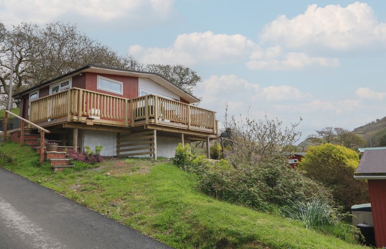 A raised red cabin with a wooden deck on a grassy slope near a road at Y Goedlan in Tywyn
