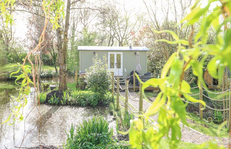 A small green building near a pond with a gravel path and wooden fence at Damsel Shepherds Hut in Badwell Ash