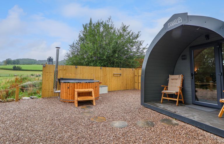An outdoor area with a wooden hot tub a gravel ground stepping stones a pod with chairs and a wooden fence at Heron Pod in Nantmawr near Trefonen