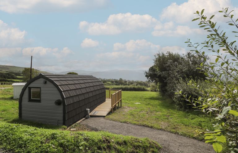 A small curved roof cabin on a grassy field with trees and hills in the background at Tarrenhendre Pod Llanegryn near Tywyn