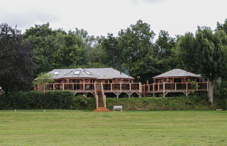 A log cabin with a wooden deck and stairs surrounded by trees and grass at Copper Lodge in Chulmleigh