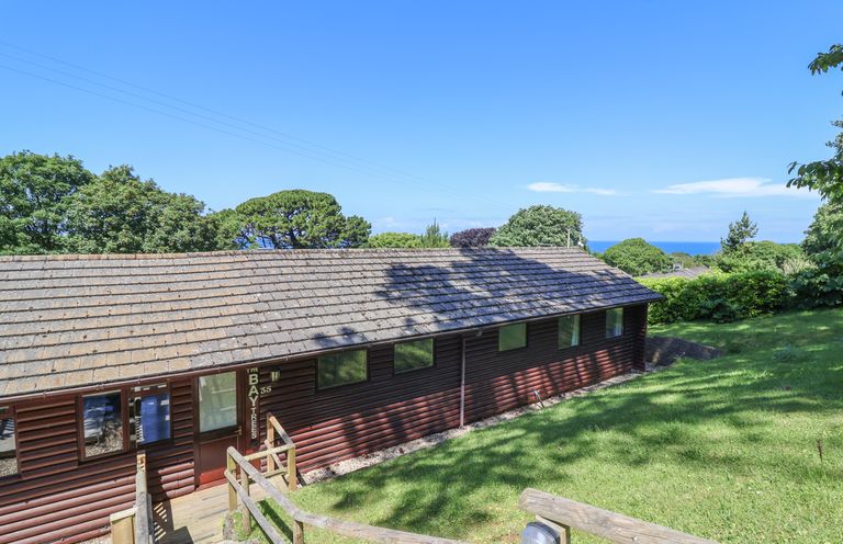 A wooden lodge with windows on a grassy slope overlooking trees and the sea at Bay Tree Lodge 35 Watermouth near Berrynarbor