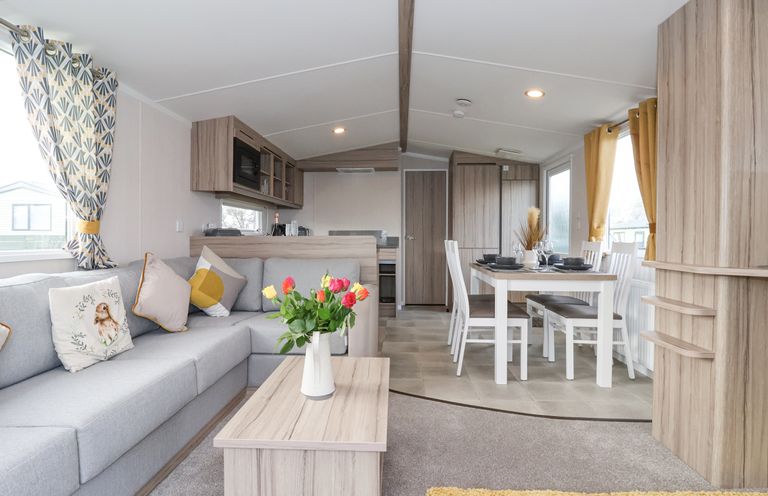 An open plan living room and dining area with a grey sofa and pillows a wooden coffee table with flowers and a dining table with chairs at Lazy Dayz Lodge East Heslerton near Malton