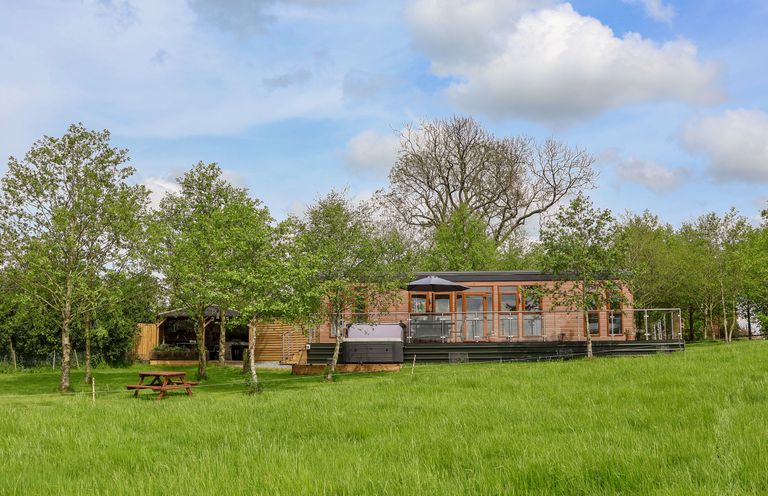 A wooden house with a deck, an umbrella, a hot tub, trees, and a picnic bench on grass at Holmside in Audley Brow near Market Drayton