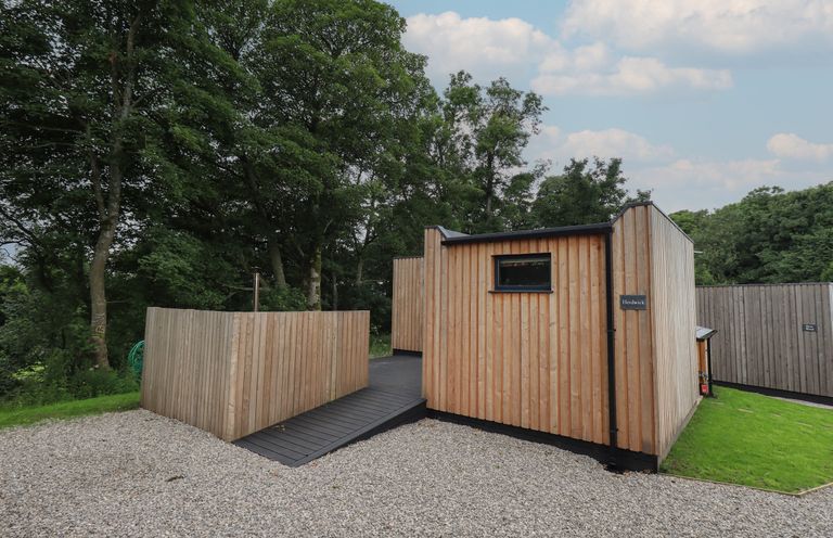 A wooden cabin with a gravel driveway and trees in the background at Herdwick Cabin in Hutton John near Penruddock
