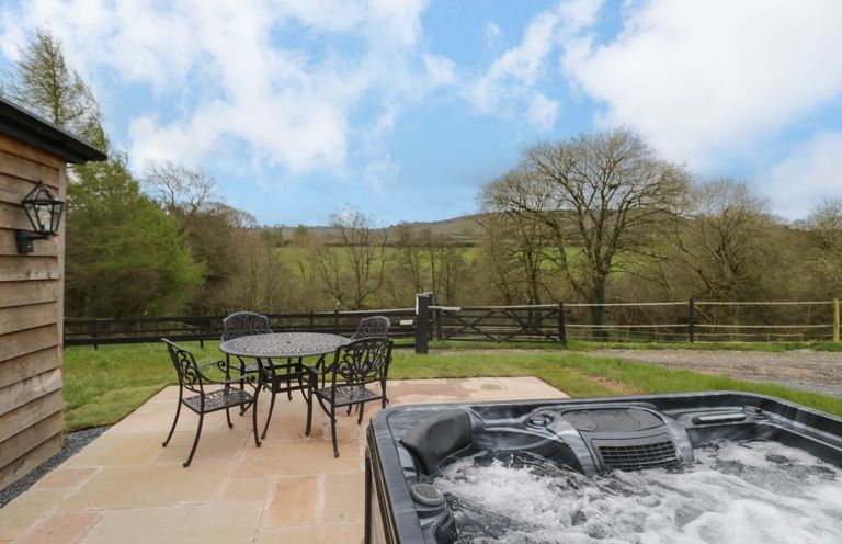 An outdoor patio with a round metal table and four chairs next to a bubbling hot tub overlooking trees and fields at Goldthorne Lodge in Clee Hill