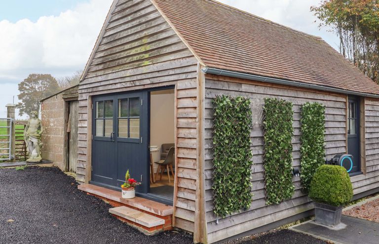 A small wooden building with a double door open showing a table and chairs inside and plants on the exterior walls at Kingfisher Cottage in Ottinge near Lyminge