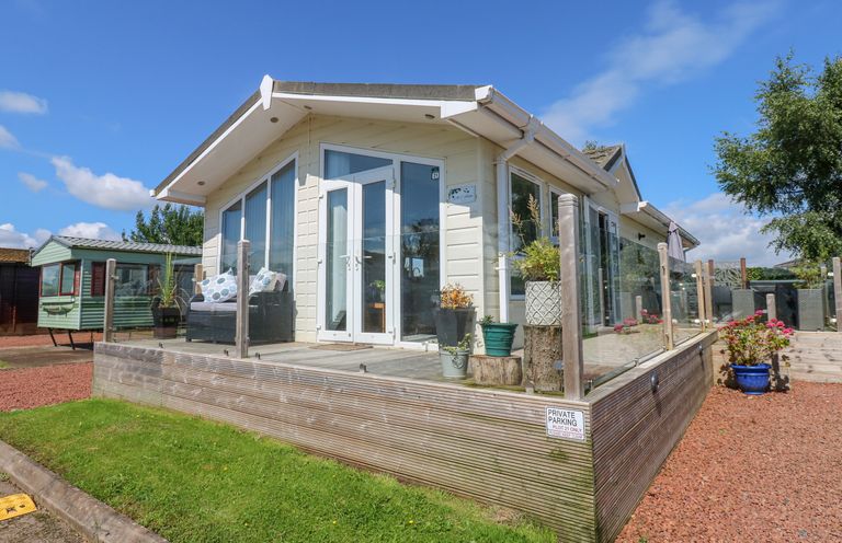 A small house with a wooden deck and glass railing surrounded by grass and gravel at A and C Lodge-Plot 21 in Armathwaite
