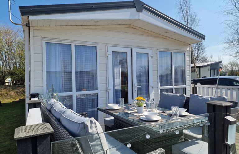 An outdoor patio area with wicker furniture and glass table set with dishes outside a small building at Ladybird Lodge South Lakes Leisure Park near Carnforth