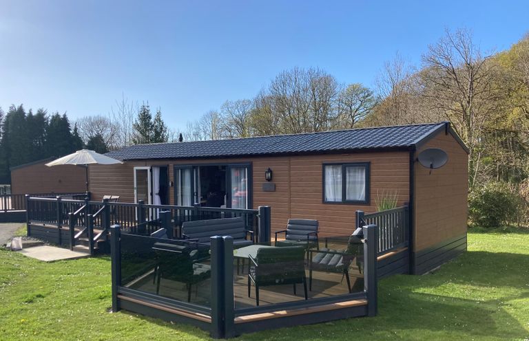 A wooden cabin with black railing deck chairs and a table on a grassy area at 43 The Deri in Llanarth