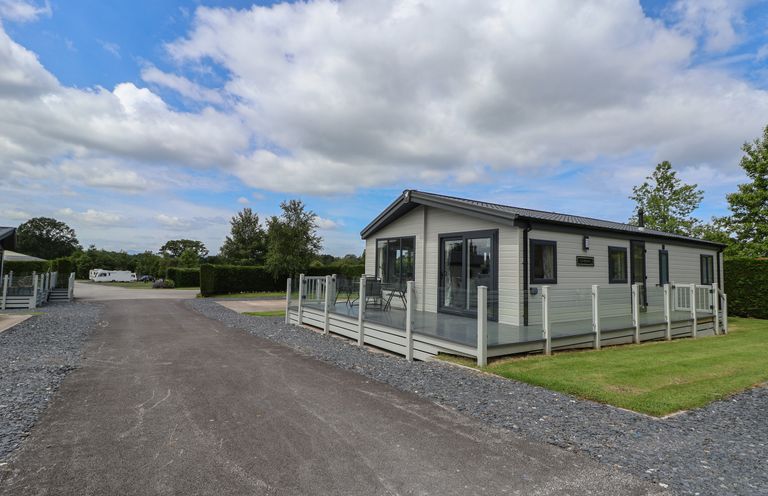 A single-story prefabricated house with a glass railing and a patio on a grassy plot by a paved road at Lodge 3 in St Asaph