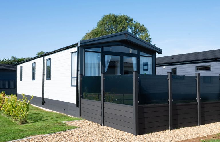 A modern static caravan with black trim and a glass panel fence on a gravel pathway surrounded by grass at Tiggywinkles in Chipping Norton