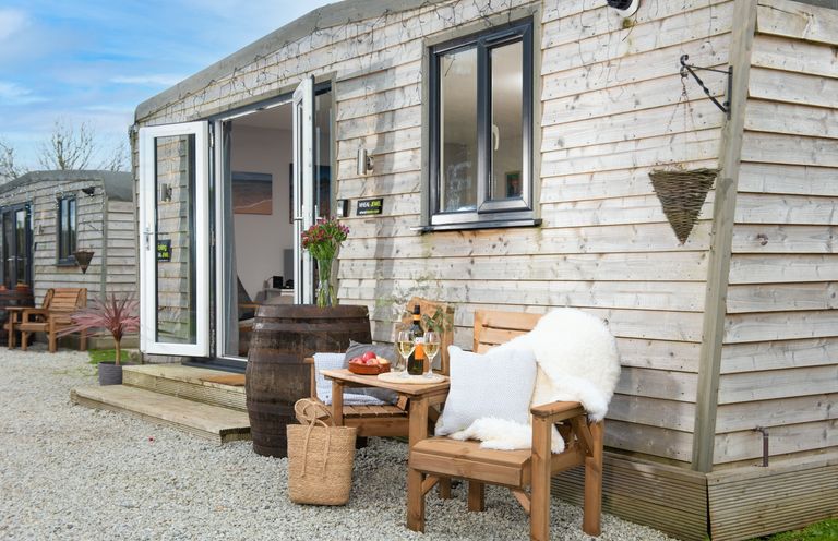 An outdoor seating area with wooden chairs and a small table with drinks outside a wooden cabin at Wheal Jewel Lodge in Wendron near Helston