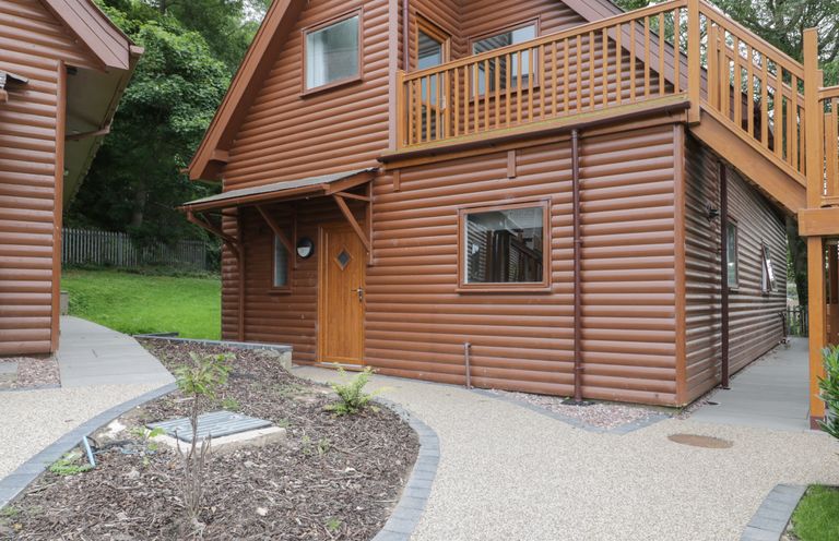 An exterior view of a wooden lodge with a door window balcony and pathways surrounded by plants and trees at Classic Lodge 25 in Dyserth