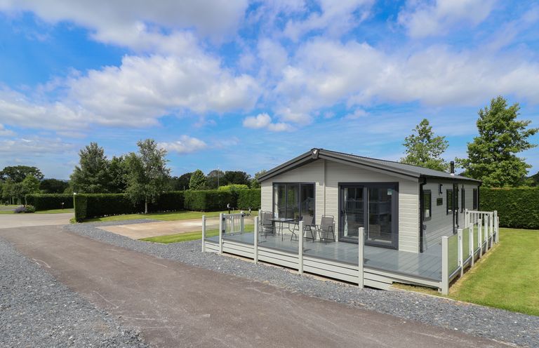 A single story house with a glass enclosed porch on a paved driveway with trees and hedges surrounding it at Lodge 9 in St Asaph