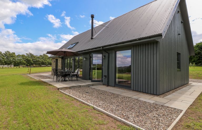 A house with a gray metal roof and dark vertical siding with an outdoor table umbrella and chairs on a patio at Strawberry Cottage in Wester Essendy near Dunkeld