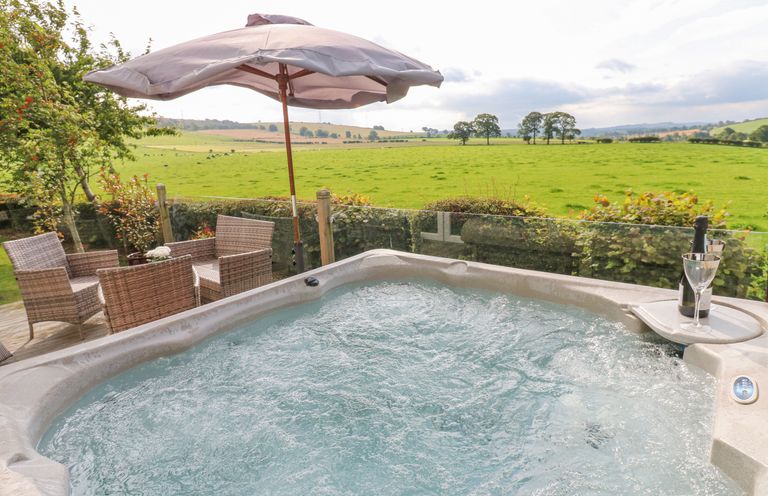 An outdoor hot tub with bubbling water next to a patio table with four chairs and a large umbrella overlooking a green field at Plot 32 High Hesket near Armathwaite
