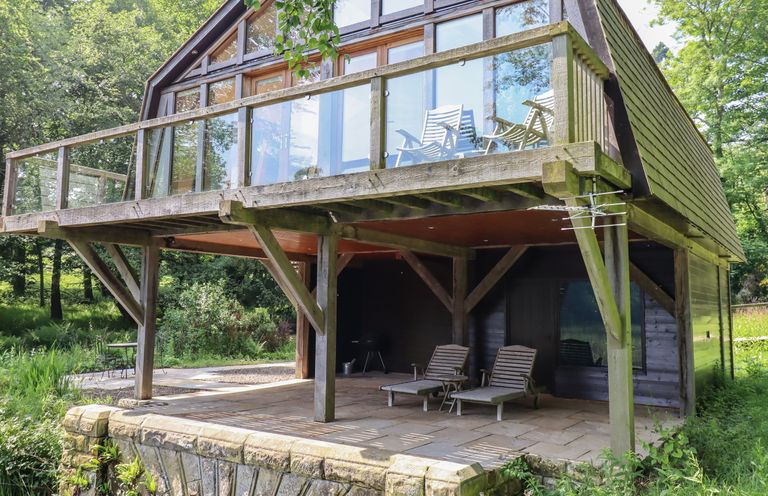 A wooden house with a balcony and outdoor chairs surrounded by trees at The Boathouse in Belford