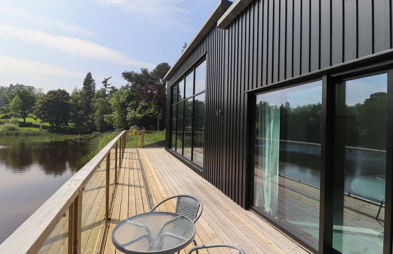 A wooden deck with glass railing and a metal table with two chairs outside a black wooden building overlooking a body of water at Otters Cove in Belford