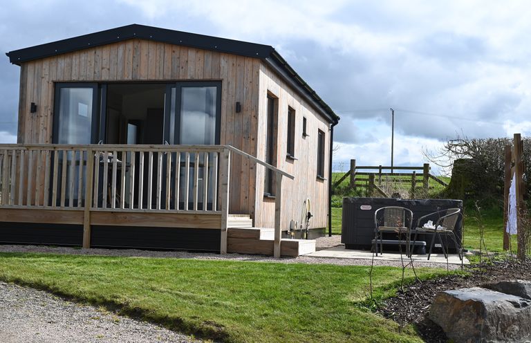 An exterior view of a wooden cabin with a small porch and railing a hot tub with two chairs next to it on a patio at Cae Pren in New Mills near Llanfair Caereinion