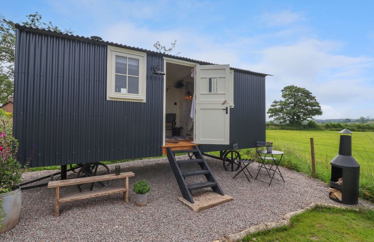 A small black corrugated metal shepherd hut with open door on a gravel area with bench table and chairs and fire pit at La'al Ling in Penrith