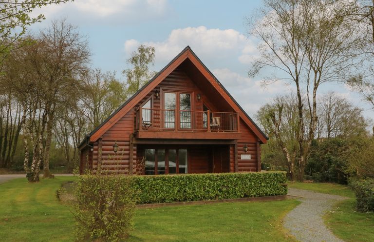 A wooden cabin with a balcony chairs and a hedge in front surrounded by grass and trees at Maple in Dolton