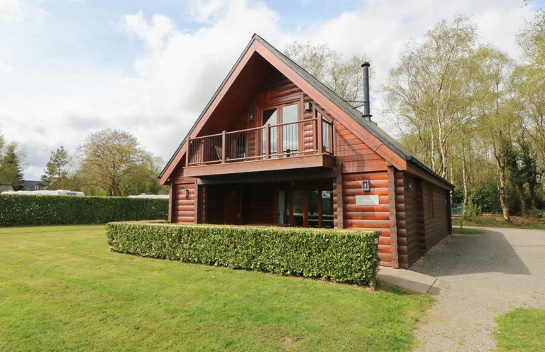 A wooden cabin with a balcony and hedge in front surrounded by grass and trees at Pine in Dolton