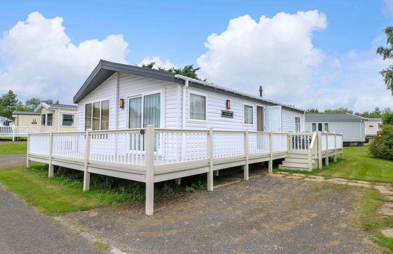 A white mobile home with a wraparound deck and steps on a grassy plot at 21 Norman Drive Lodge in Burgh Castle