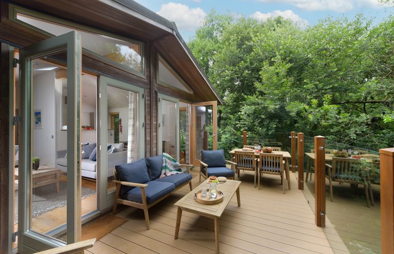 A wooden deck with blue cushioned chairs a wooden table and glass railing surrounded by trees at 10 Stonerush Valley Lanreath near Looe