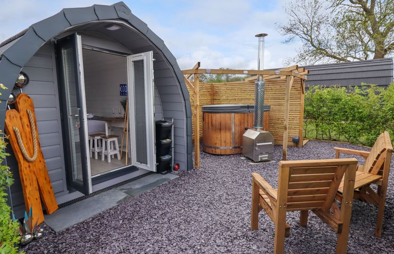 An outdoor area with two wooden chairs a wooden hot tub a gray pod-shaped cabin with open doors at Moel Famau Bryniau Clwyd near Denbigh