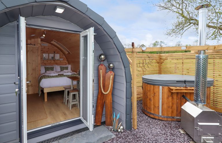 A small wooden pod with open doors showing a bed inside and a covered hot tub with heater outside at Moel Arthur in Bryniau Clwyd near Denbigh