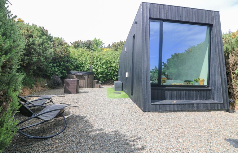 A gravel patio area with two black curved lounge chairs a black wooden building with a large window and a seating area and a hot tub surrounded by green bushes at Sunriser in Tara Hill near Ballymoney County Wexford