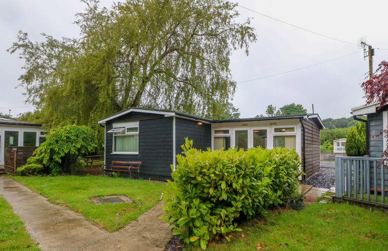 A small dark wooden house with white framed windows and a pathway leading to the entrance surrounded by greenery and trees at 245 Riverside Lodge in Caeathro