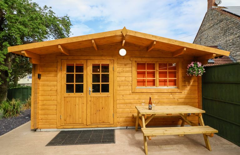 A wooden cabin with double doors and a window next to a wooden picnic table with a bottle and two glasses on it at Log Cabin - Amber Glow in Glastonbury