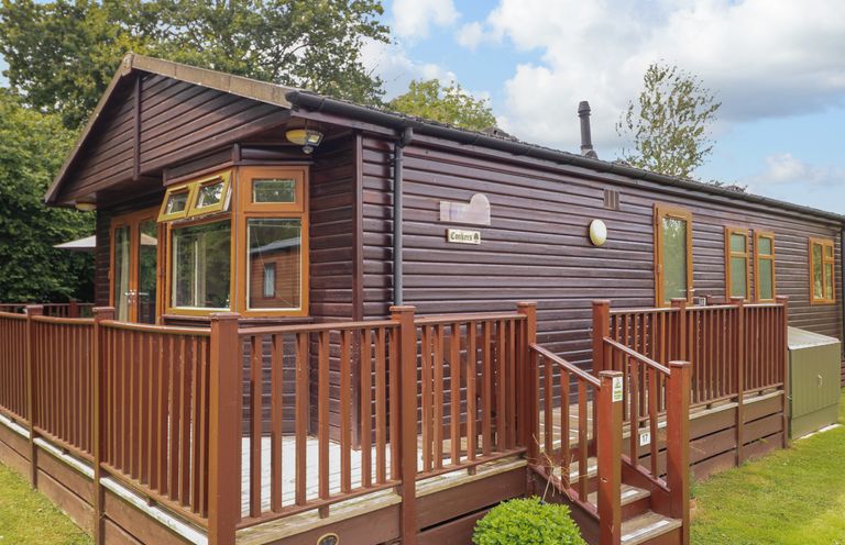An exterior view of a wooden lodge with a porch and railing surrounded by grass and trees at The Brambles 17 - Conkers Lodge Finlakes Holiday Park near Chudleigh