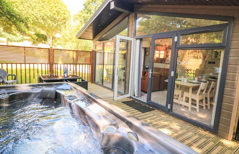 An outdoor deck with a hot tub a table with chairs and a dining area visible through glass doors at Woodland View 7 - Oak Cabin Finlakes Holiday Park near Chudleigh