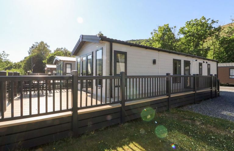 An exterior view of a white mobile home with a wooden deck and black railing surrounded by grass and trees at Saorsa in St Fillans
