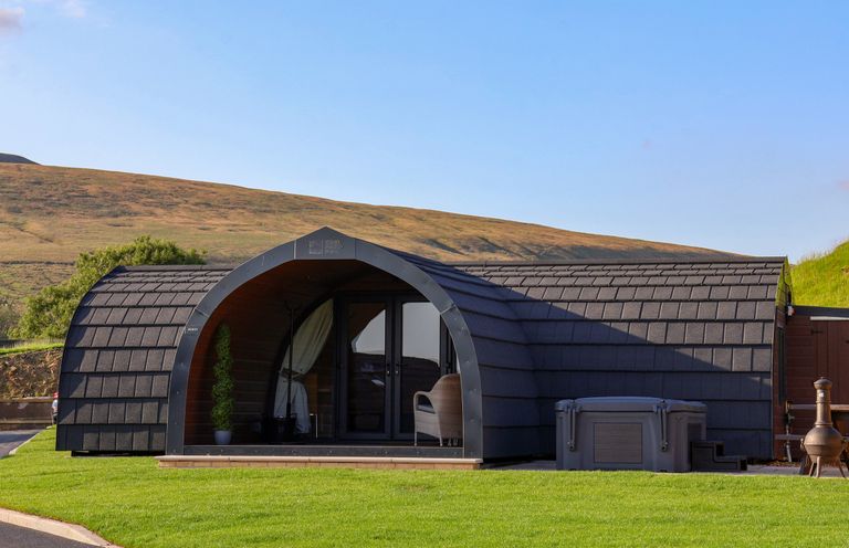 A curved black cabin with glass doors and a wicker chair outside on green grass at Heron in Ingleton