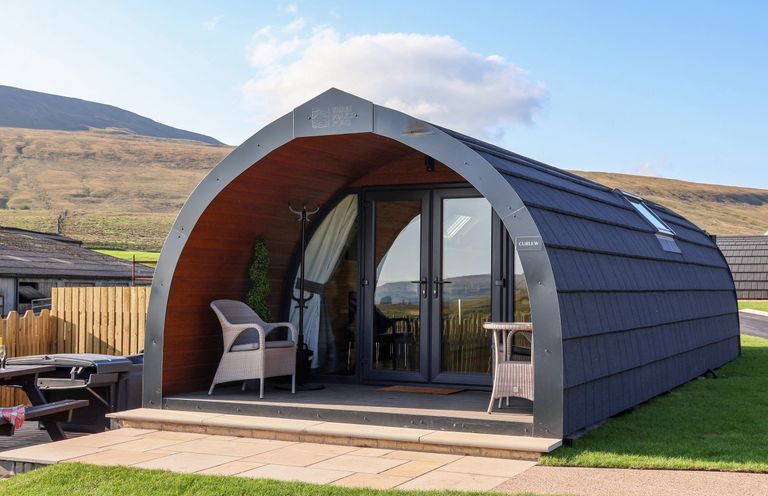 A curved wooden pod with glass doors, two wicker chairs, and a small table on the porch at Curlew in Chapel Le Dale near Ingleton