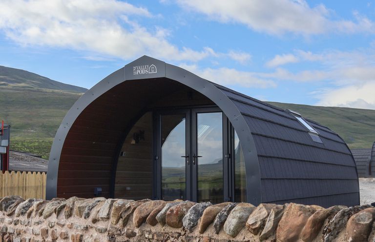An arched pod cabin with glass doors behind a stone wall in a rural landscape at Woodpecker in Chapel Le Dale near Ingleton