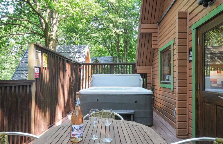An outdoor wooden deck with a hot tub and a table with a bottle and two wine glasses at Lodge Nine Finlake Holiday Park near Chudleigh